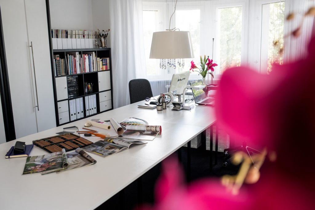 Ein Arbeitszimmer in Pink mit Arbeitstisch, Lampe, Schrank und Regal hinter pinkfarbenen Blüten