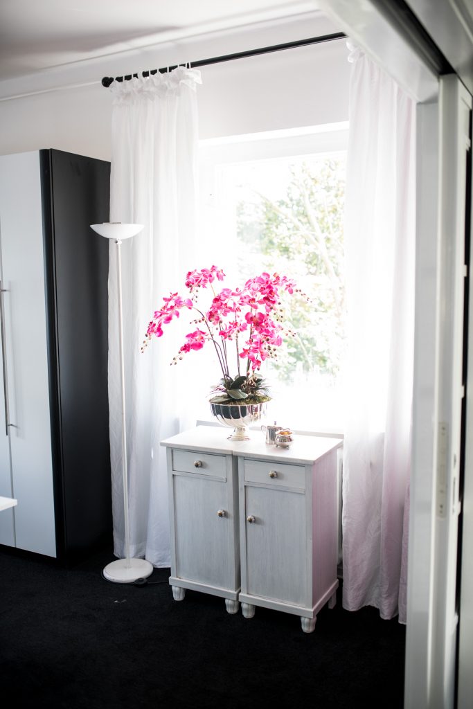 Ein Arbeitszimmer in Pink mit pinkfarbenen Orchideen auf kleinen weißen Schränkchen vor dem Fenster