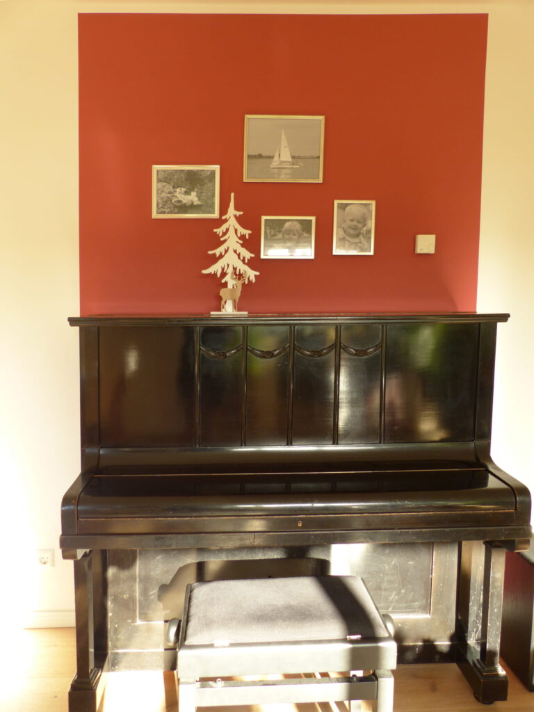 altes Klavier nach Umgestaltung mit Klavierhocker und weissen Bilderrahmen vor rotem Farbspiegel an der Wand
