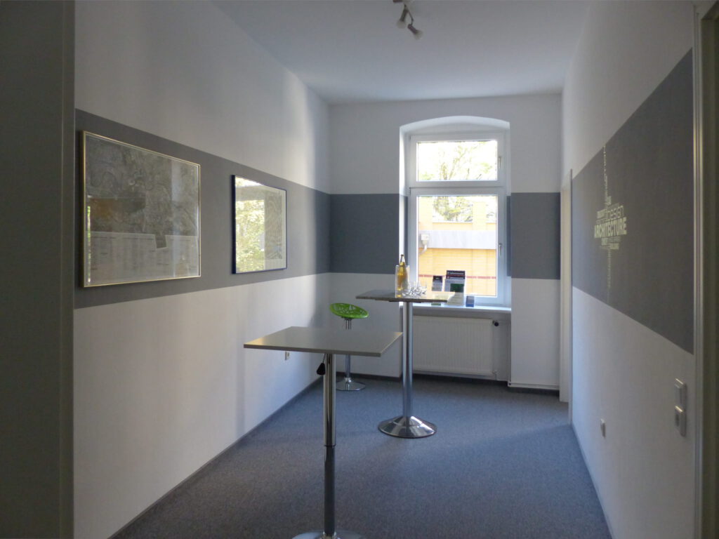 Schulungsraum nach Umgestaltung mit Fenster und Bildern in weißer Wand und grauerm Farbstreifen und Stehtischen auf grauem Teppich