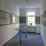 Schulungsraum nach Umgestaltung mit Fenster und Bildern in weißer Wand und grauerm Farbstreifen und Stehtischen auf grauem Teppich