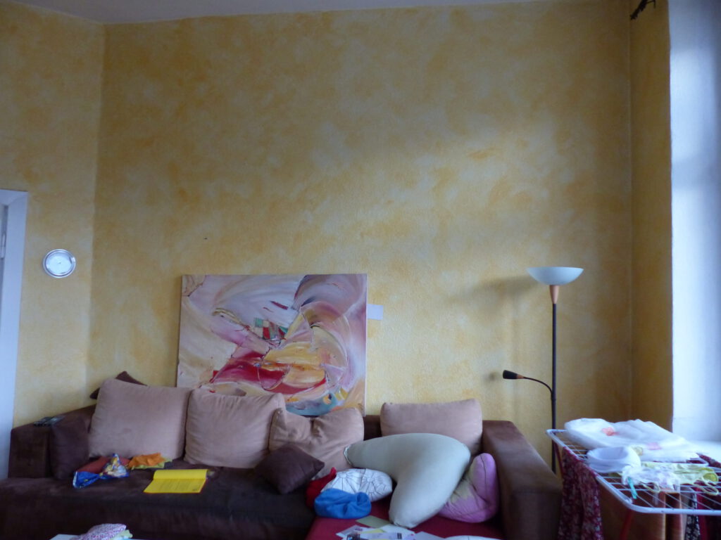 Wohnzimmer Blick 5 vorher mit Ecksofa und Waeschestaender vor gelber Wand